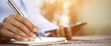 Image of the hands of a black woman writing in a notebook while holding a cell phone