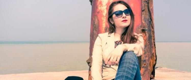 Photo of a pensive young woman leaning against a rusty post, near the ocean. She has a bag and laptop sitting next to her.