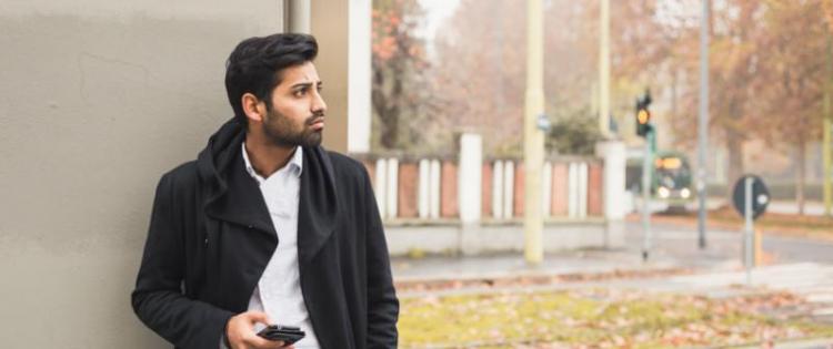 Photo of a man holding his phone and leaning against a wall in a neighborhood. 