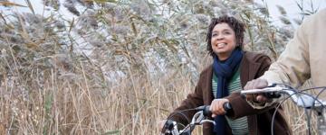 Photo of a woman on a bicycle in autumn, she wears a coat and scarf. A second cyclist can be partially seen to her side.