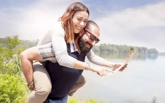 Photo of a smiling couple by a lake, a man holds a woman on his back while she holds out a map for them both to see.